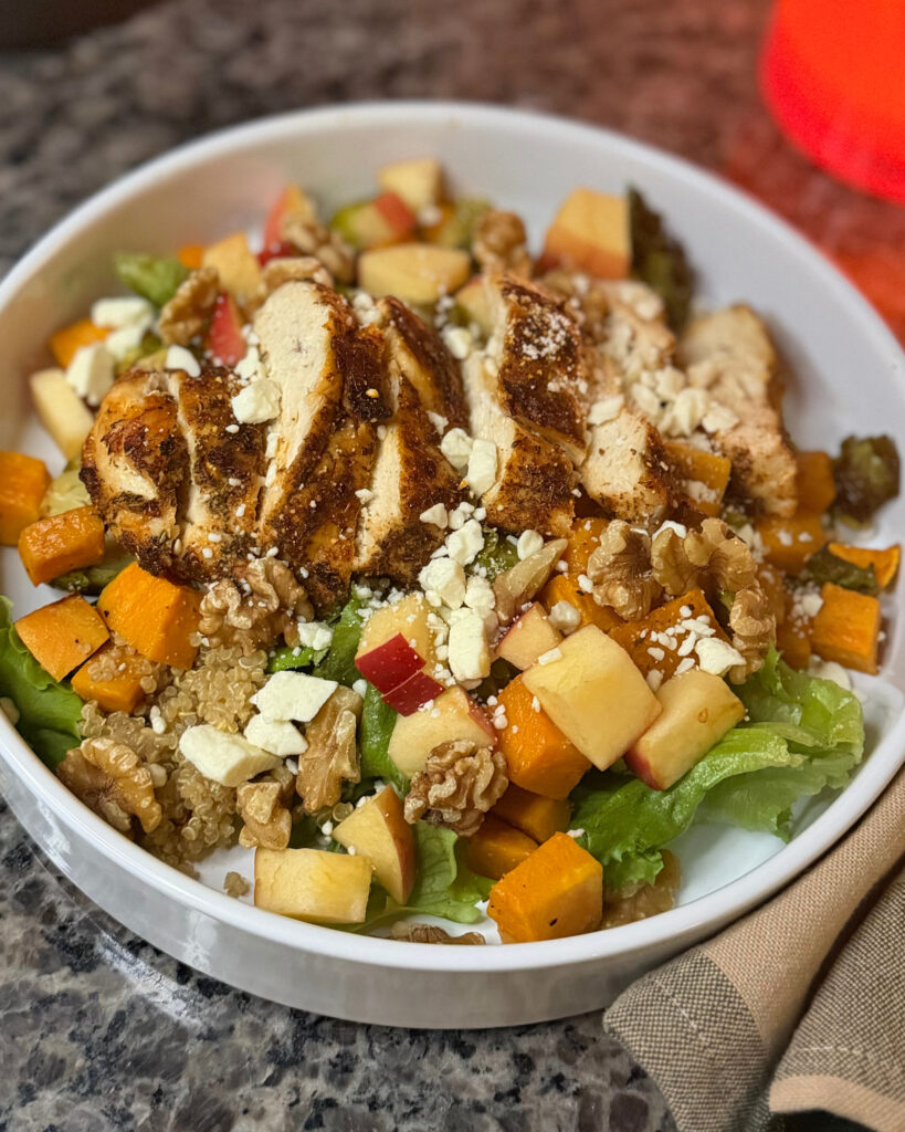 Close-up of sliced seasoned chicken over quinoa, greens, sweet potatoes, apple, walnuts, and cheese