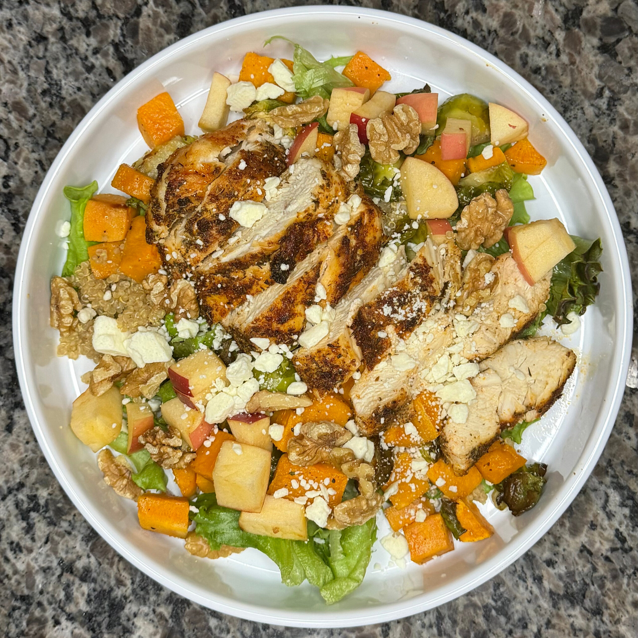 Overhead view of a chicken harvest bowl with quinoa, roasted vegetables, apple, walnuts, and cheese